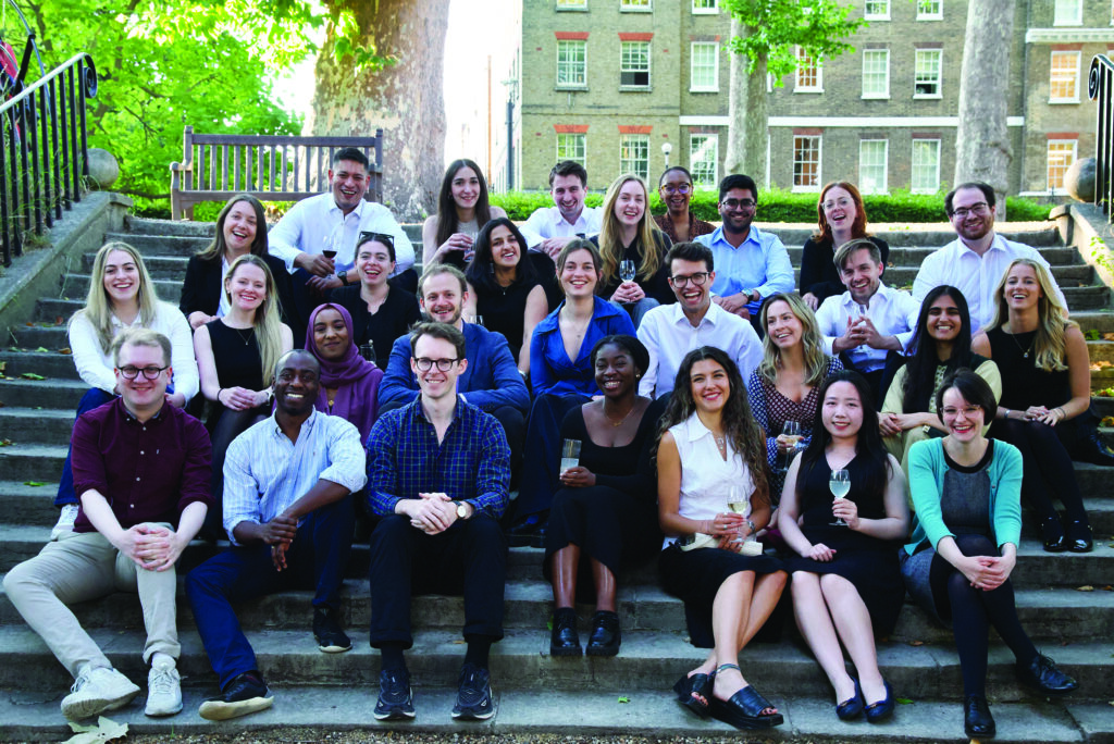 Group photo of the Inn’s residential scholars sitting on outdoor steps. The diverse group of men and women are smiling and holding drinks, dressed in smart-casual attire, with a bench and greenery visible in the background