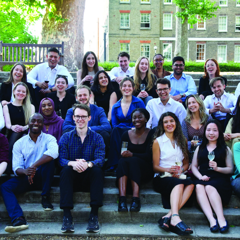 Group photo of the Inn’s residential scholars sitting on outdoor steps. The diverse group of men and women are smiling and holding drinks, dressed in smart-casual attire, with a bench and greenery visible in the background