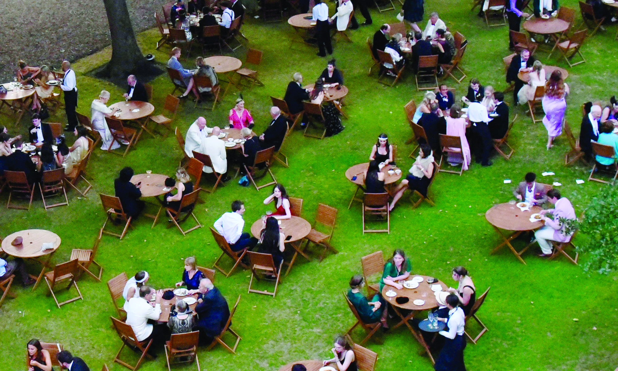 Arial picture of people at tables on the grass during an evening party event with trees and string lights in the background