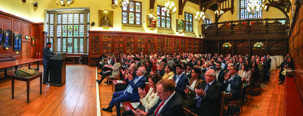 A speaker addresses a full audience in an elegant, wood-paneled hall with stained glass windows. 