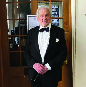 Master Lord Beloff, stood with hands crossed over each other on his front, wearing a suit and bow-tie, in front of a wooden door with window panelling.