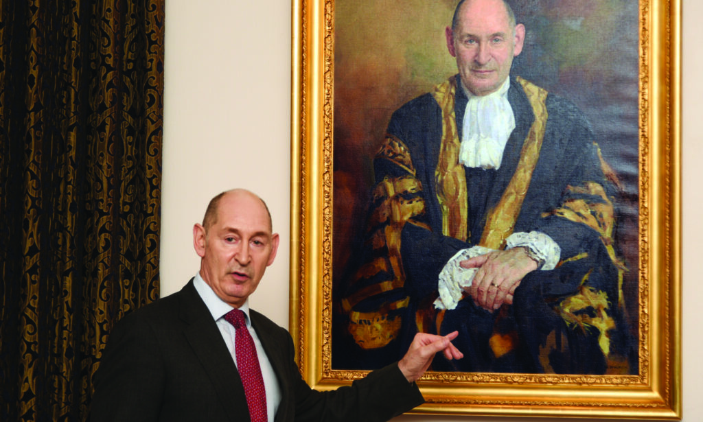 Late Master Terence Etherton stood with his arm towards his portrait which has a painting of him in his robes, with a gold frame. Master Etherton is wearing a suit and red tie.