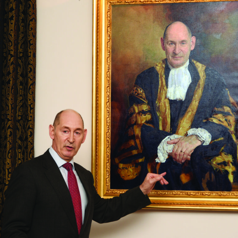 Late Master Terence Etherton stood with his arm towards his portrait which has a painting of him in his robes, with a gold frame. Master Etherton is wearing a suit and red tie.
