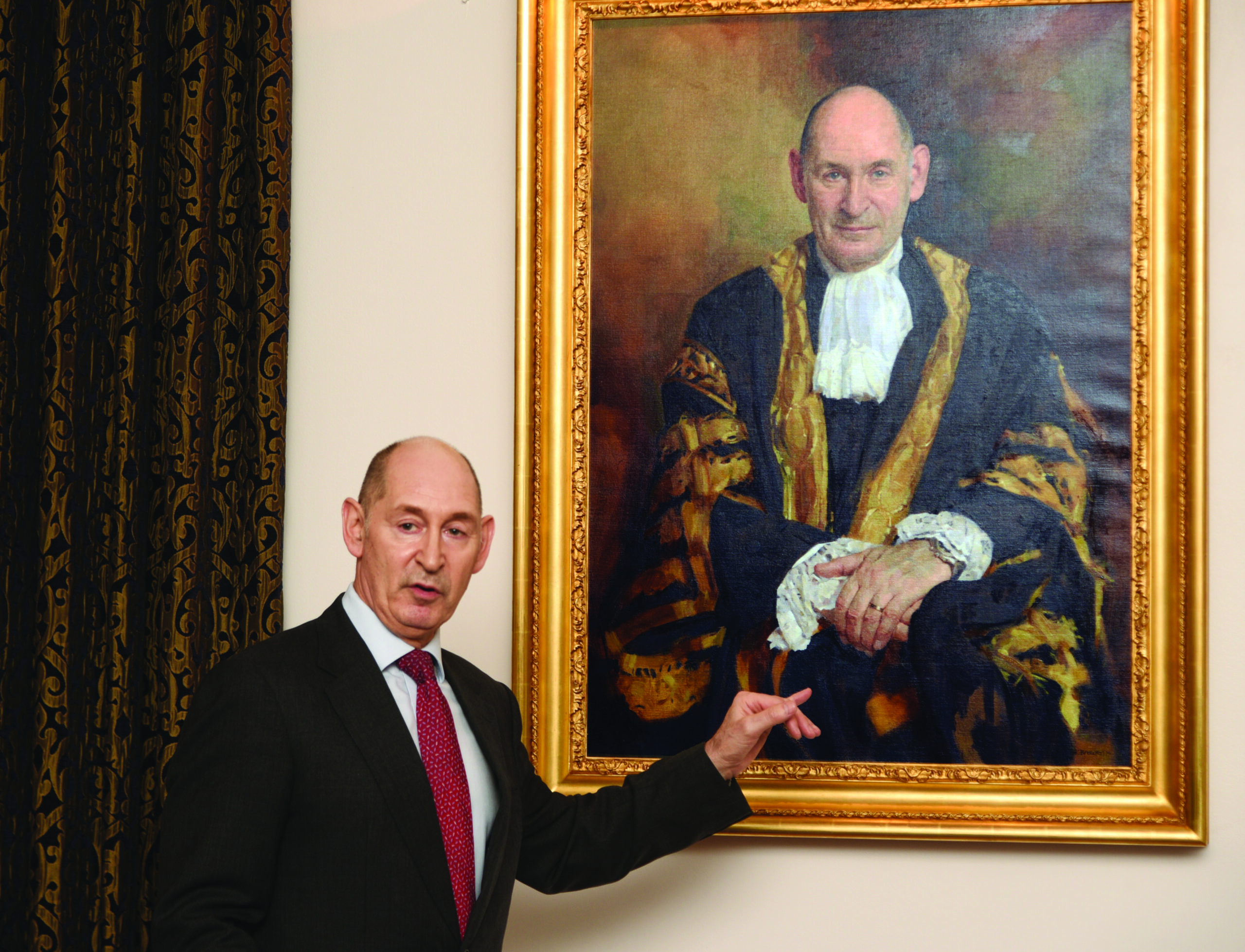 Late Master Terence Etherton stood with his arm towards his portrait which has a painting of him in his robes, with a gold frame. Master Etherton is wearing a suit and red tie.