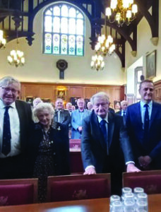 A group of elderly people stands smiling in a warmly lit Hall with wooden pews and chandeliers. A stained glass window is visible in the background.