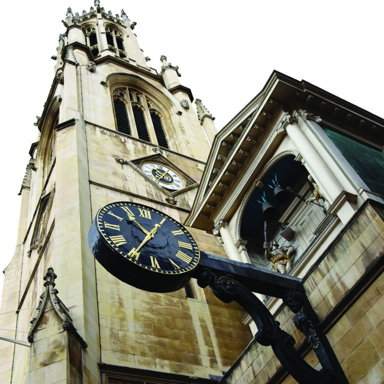 Tall stone church tower with ornate details rises against a cloudy sky. A black clock with gold Roman numerals hangs prominently in the foreground.