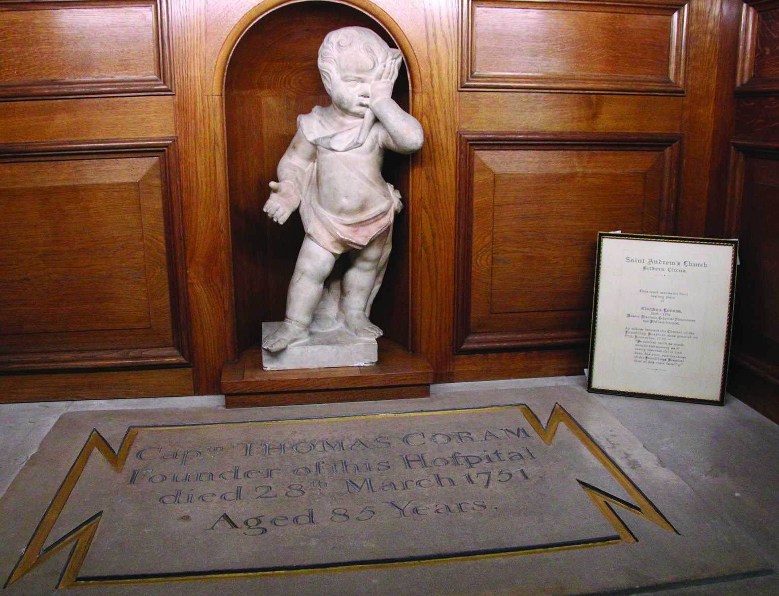 A marble cherub statue stands against a wooden wall, with a commemorative plaque nearby. A stone slab with inscriptions lies in the foreground