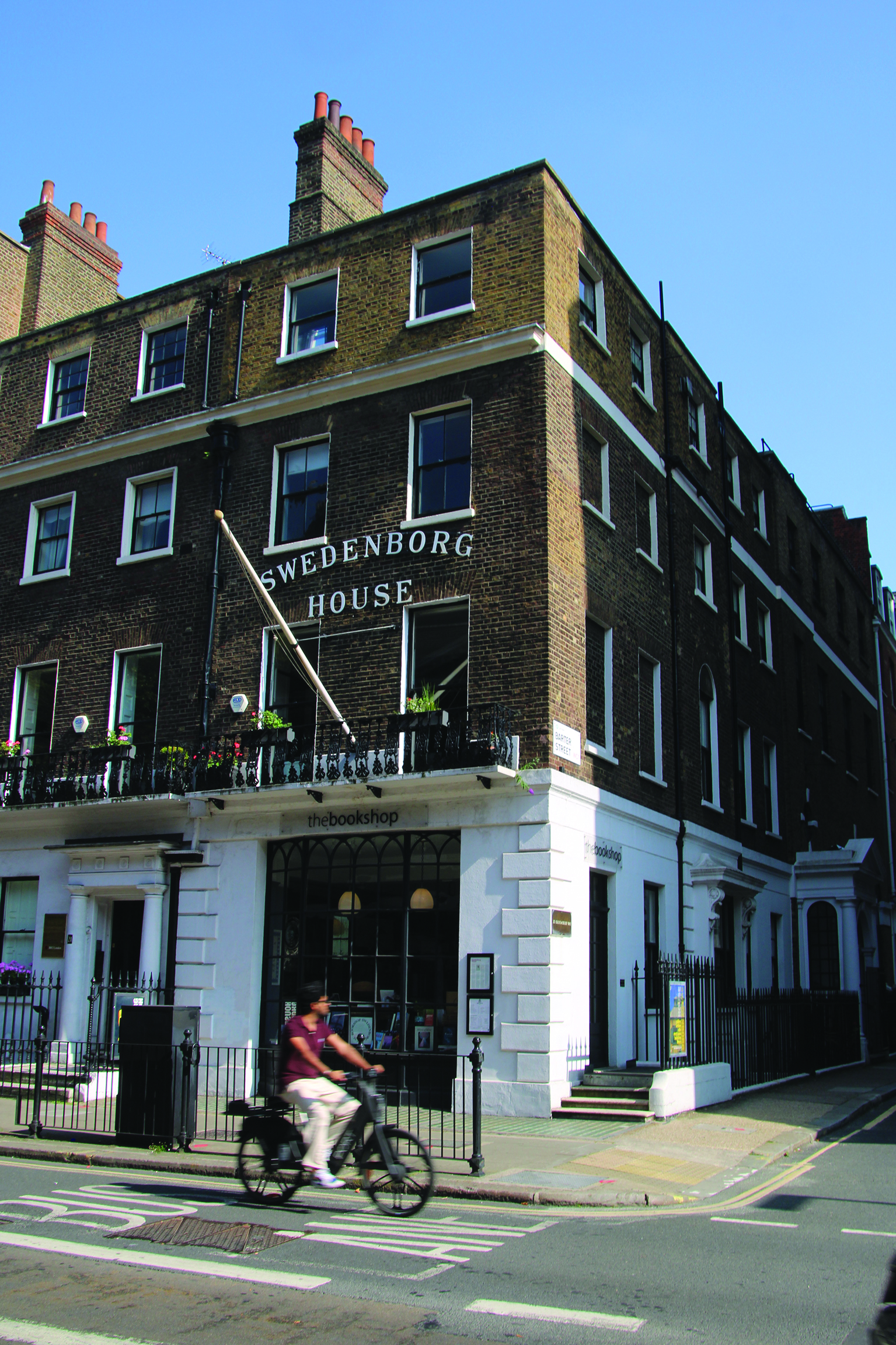 Street view of Swedenborg House, a four-story brick building with white trim, featuring chimneys and sash windows. A person on a bicycle passes by.