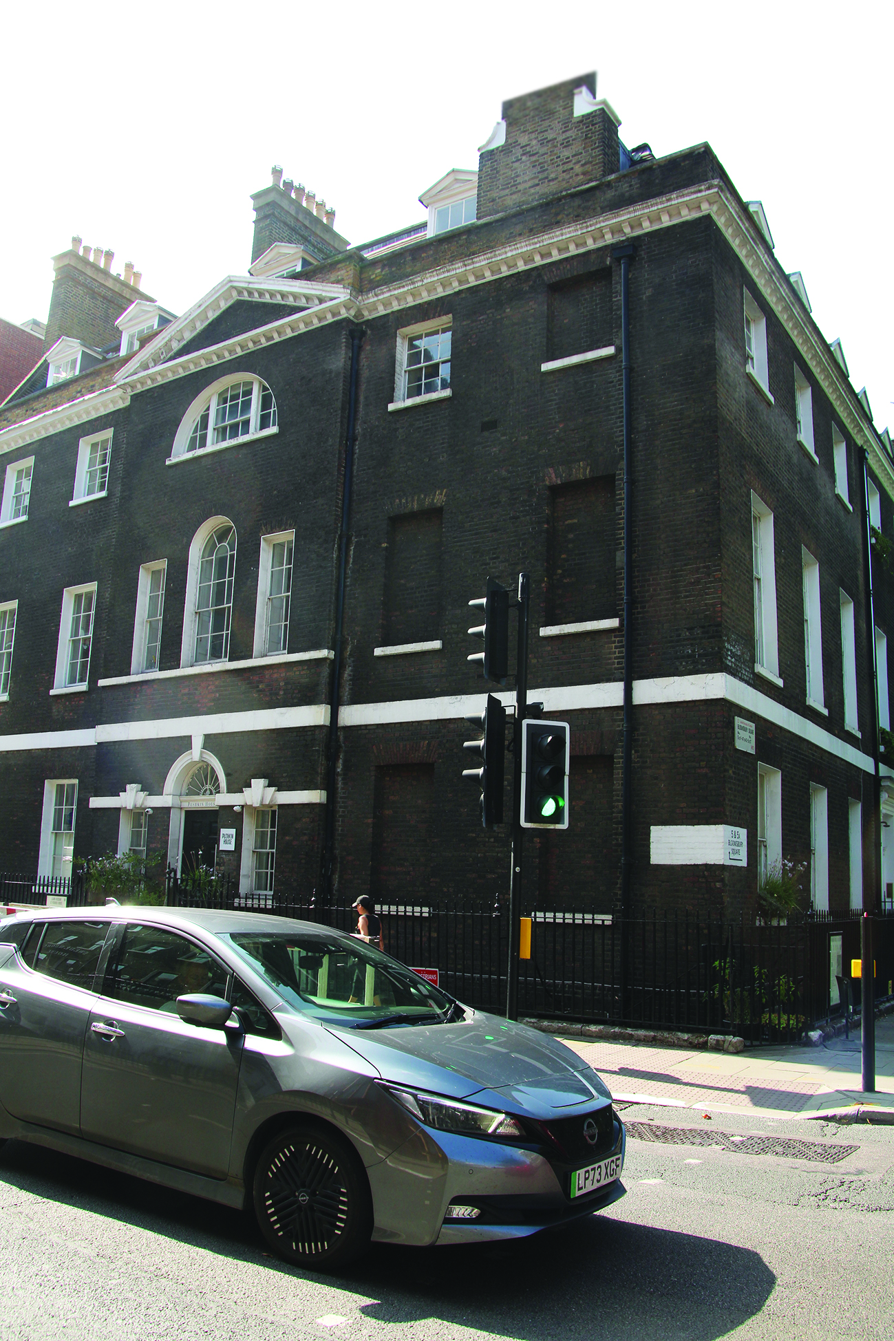 A brick building with white trim features arched and rectangular windows. A green traffic light is in front, and a person walks near a car.