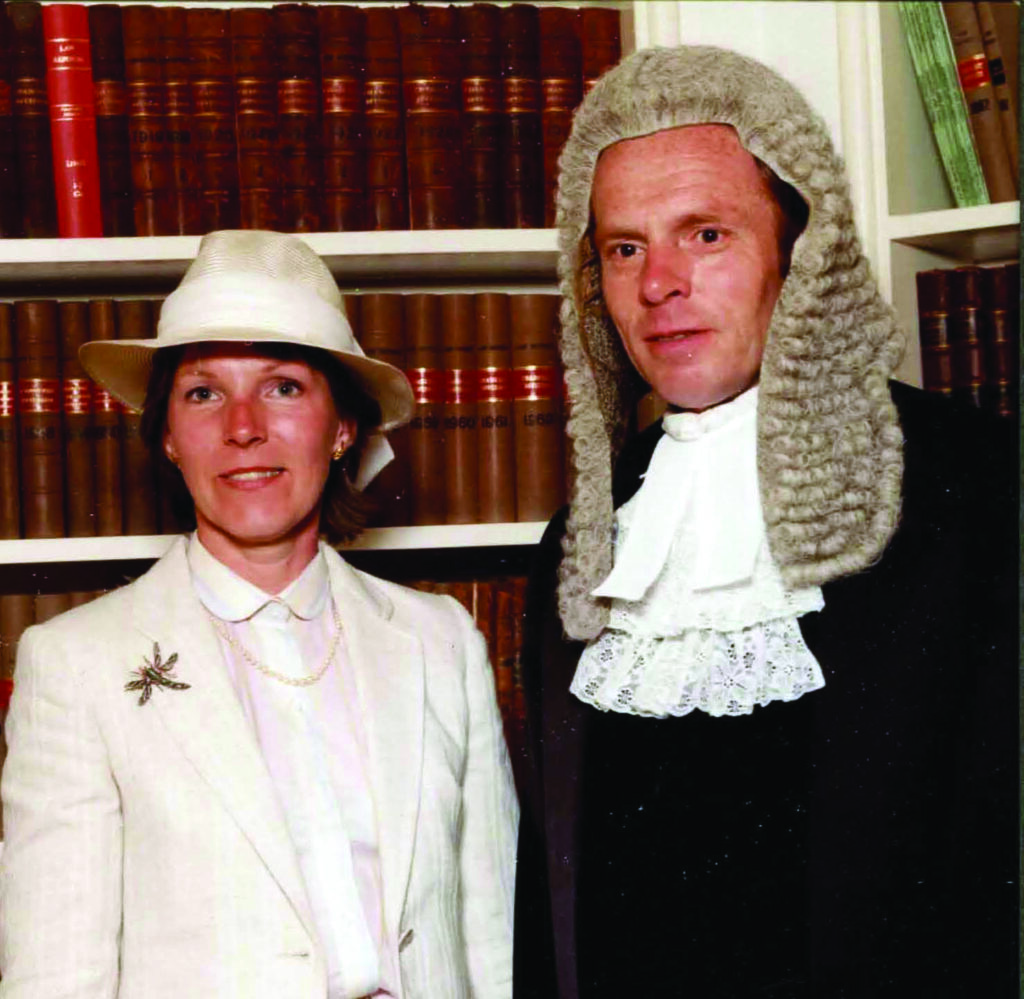 A woman in a white suit and hat stands beside Master Richard Hawkins in a judge's wig and robe. They are in front of a bookshelf filled with leather-bound book