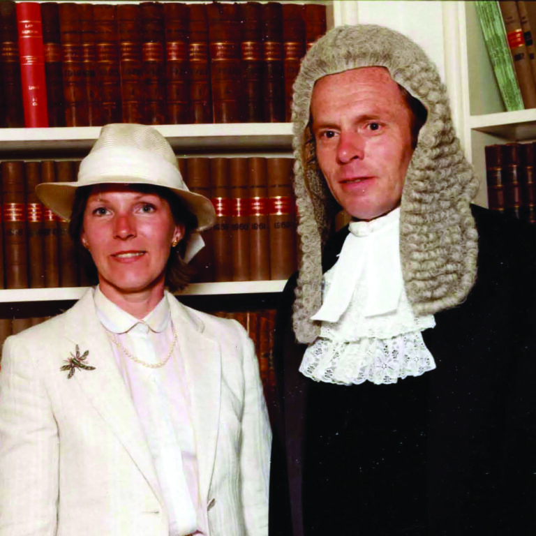 A woman in a white suit and hat stands beside Master Richard Hawkins in a judge's wig and robe. They are in front of a bookshelf filled with leather-bound book