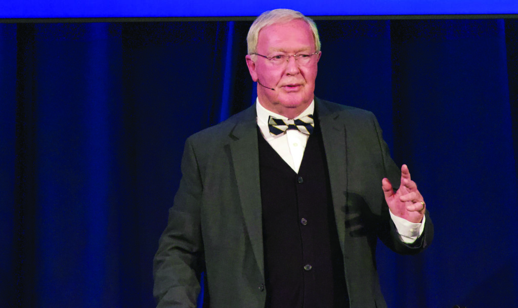 Speaker in a grey suit wearing a bow tie and glasses, against a royal blue background