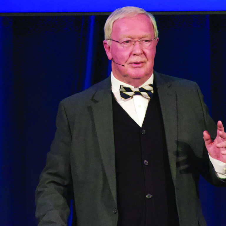 Speaker in a grey suit wearing a bow tie and glasses, against a royal blue background