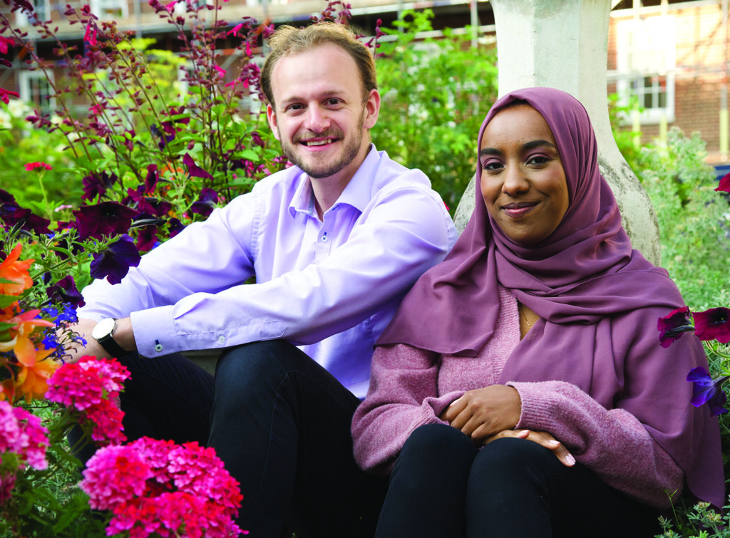 Two residential scholars sit in a garden with vibrant flowers. The man wears a light purple shirt, and the woman wears a pink hijab and sweater, both smiling softly.
