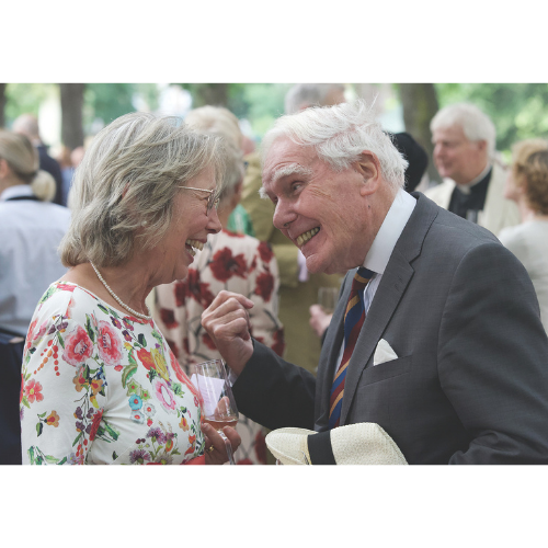 Two members in a crowded garden party. A woman on the left wears a white and pink floral dress and smiles to the man on the right. The man on the right wears a dark suit, colourful tie and carries a light coloured summer hat.