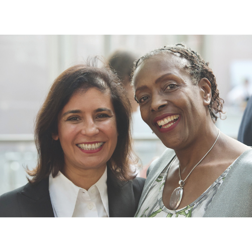 Two women smiling to camera. On the left, with shoulder length dark hair, a black jacket and white shirt. On the right, short hair, a floral top and a large silver pendant.