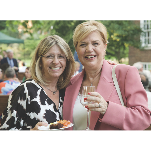 Two members smiling with a glass of champagne and a small plate of food in the Inn's gardens. The woman on the left has shoulder length blond hair, glasses, and wears a black and white top. The woman on the right has short blond hair and wears a light pink jacket.
