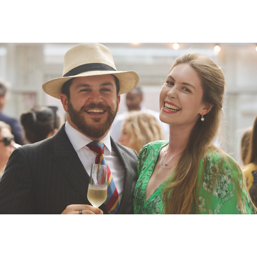 Two members smiling with a glass of champagne. The man on the left has a short beard and wears a cream summer hat, suit and colourful tie. The woman on the right wears a green floral dress and has long brown hair.