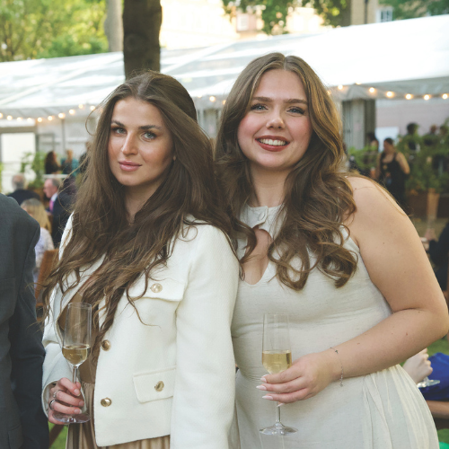 Two members smiling and holding glasses of champagne. They have long dark hair. One is wearing an off-white dress. The other a light coloured jacket.