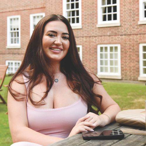 Woman seated at a table in the gardens, with a red brick building with white windows in the background. She has long dark hair and is wearing a light pink dress