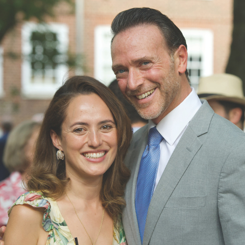 Two people smiling. A woman on the left wears a floral dress. A man on the right wears a gray suit with a blue tie.