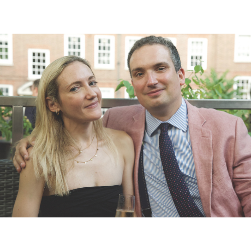 Two people smiling. A woman on the left wears a black dress. A man on the right wears a light pink suit with a blue shirt.
