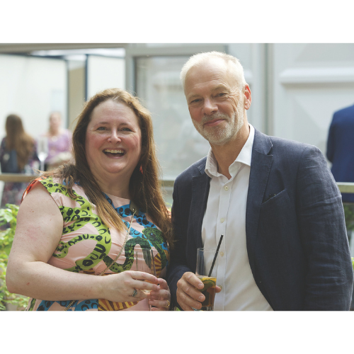 Two people smiling. On the left a woman with long hair and a colourful dress. On the right a man in a suit.