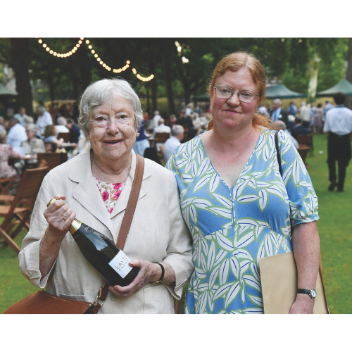 Two women at a garden party. One is holding a bottle of champagne and one a large envelope.