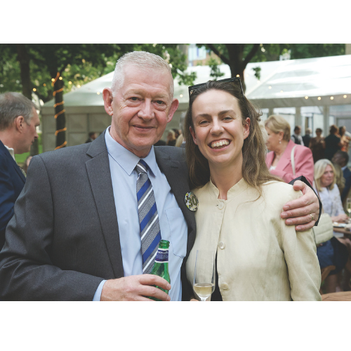 Two people smiling. A man on the left has short white hair, wears a blue suit and tie and holds a bottle of beer. The woman on the right has sunglasses on her head, wears a light coloured jacket and holds a glass of champagne.