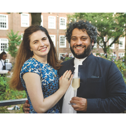 Two members smiling in the Inn's garden. The woman on the left wears a blue floral dress and has long brown hair. The man on the right has a beard and short dark hair and wears a dark suit whilst holding a glass of champagne