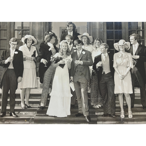 Guests and Penny and Nick Hamilton on their wedding day standing on steps, holding drinks. The bride and groom are centered, surrounded by guests in formal attire and hats, smiling brightly.
