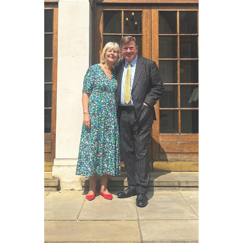 Penny and Nick Hamilton stand arm in arm outside a wooden door, Penny wears a floral dress with red shoes, and Nick is in a pinstripe suit with a yellow tie.
