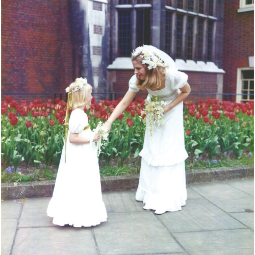 Penny Hamilton in a wedding dress and eceives flowers from a young girl in a white dress, standing outside a brick building with greenery.