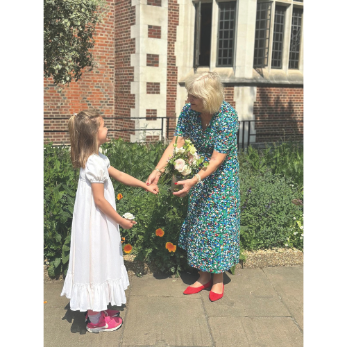 Penny Hamilton in a floral dress and red shoes receives flowers from a young girl in a white dress and pink shoes, standing outside a brick building with greenery.
