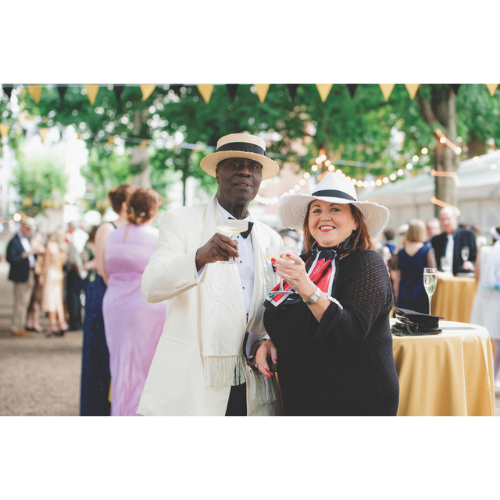 Two people smiling and raising their glasses to camera. The man on the left wears a white suite with a hat. The woman on the right wears a dark dress with a white hat.