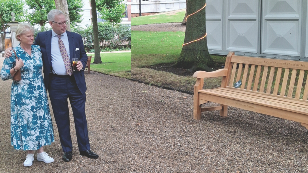 Master Richard Atkins and partner, arm in arm, glancing at the memorial bench for their late daughter, Lucy Atkins. Master Atkin wears a suit and patterned tie whilst holding a drink, his partner wears a blue and white floral dress, with a brown shoulder bag,